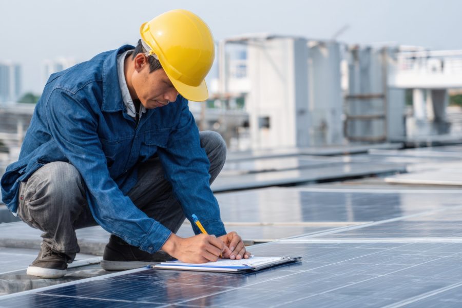 A worker in a hard hat inspects solar panels on a roof, taking notes on a clipboard. The image represents the growing renewable energy sector, technical jobs, and the installation and maintenance of green technology.