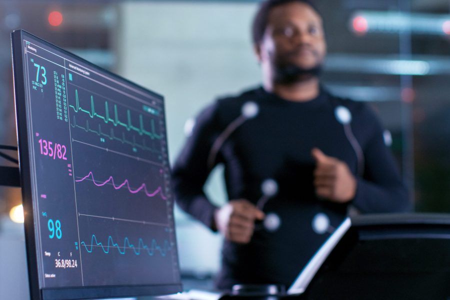 Close-up Shot of a Monitor With EKG Data. Male Athlete Runs on a Treadmill with Electrodes Attached to His Body while Sport Scientist Holds Tablet and Supervises EKG Status in the Background.