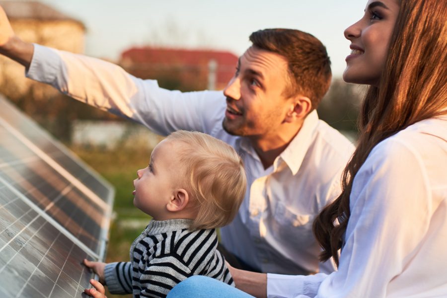 Man shows his family the solar panels on the plot near the house during a warm day. Young woman with a kid and a man in the sun rays look at the solar panels.