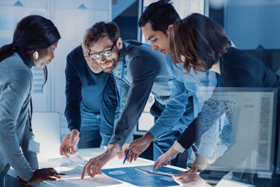 Engineers Meeting in Technology Research Laboratory: Engineers, Scientists and Developers Gathered Around Illuminated Conference Table, Talking and Finding Solution, Inspecting and Analysing Industrial Engine Design.